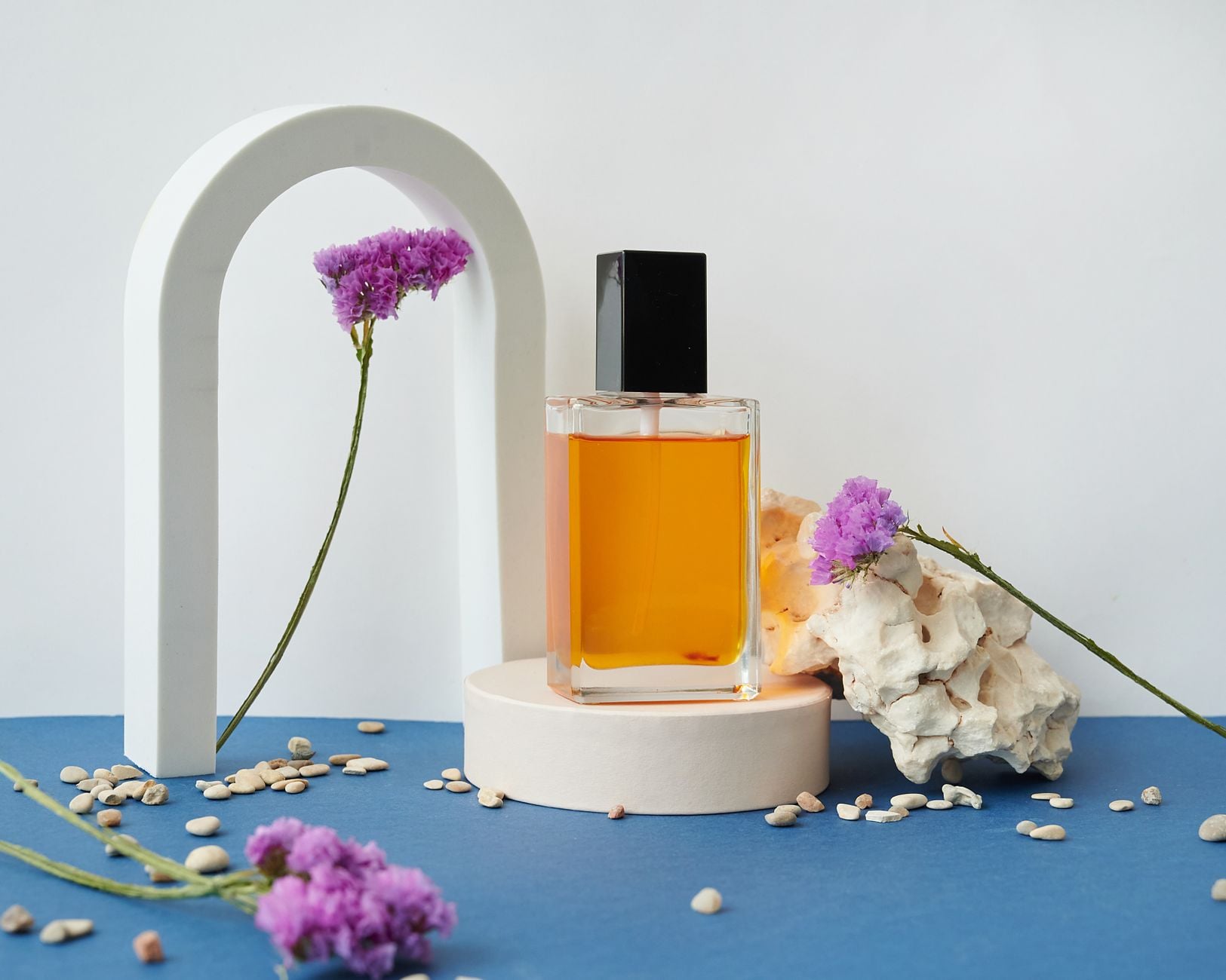 Pomarańczowe perfumy w szklanym flakonie na białym podium otoczonym fioletowymi kwiatami, białymi kamieniami i białą arką